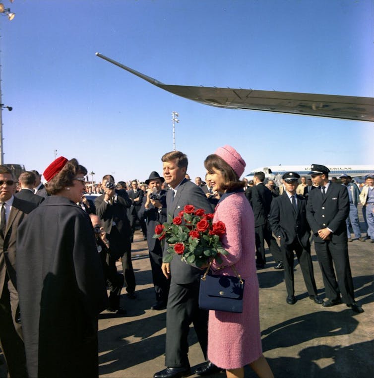 Una mujer con un traje rosa y un sombrero sostiene rosas rojas junto a un hombre con un traje gris, mientras otras personas observan.