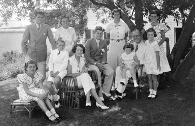 A black and white old photo shows a large family all wearing white or light colors seated together under a tree. 
