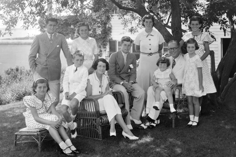 A black and white old photo shows a large family all wearing white or light colors seated together under a tree.