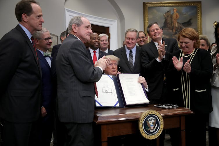 A group of standing people applaud as a man seated behind a desk bearing the U.S. presidential seal holds up a document.