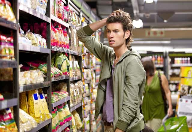 Young man looking worried scratching his had in supermarket in front of pasta shelf