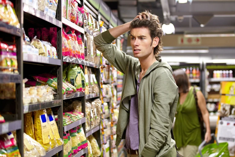 Young man looking worried scratching his had in supermarket in front of pasta shelf