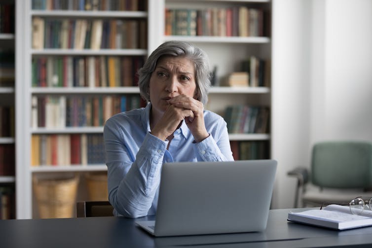 A woman sits at a desk with a laptop and bookshelves behind.