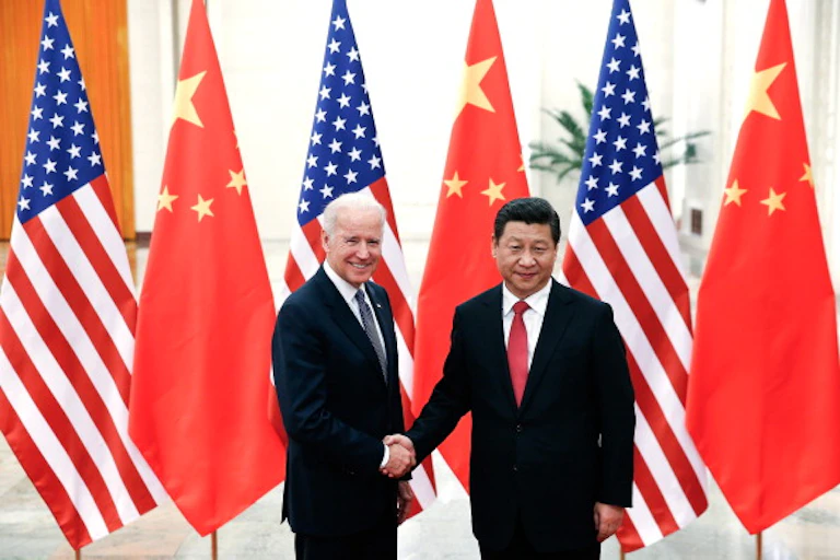 Two men shake hands in front of large flags of the United States and China.