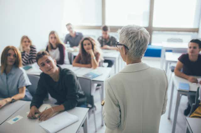 Profesora de espaldas con pelo y traje blancos, frente a un grupo de 8 alumnos sentados en sus pupitres.