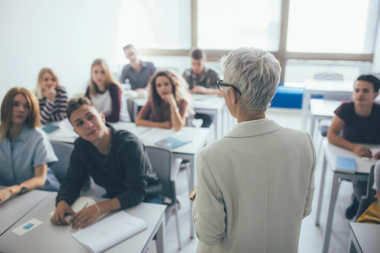 Profesora de espaldas con pelo y traje blancos, frente a un grupo de 8 alumnos sentados en sus pupitres.