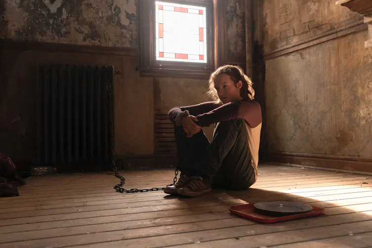 A photo of Bella Ramsey (who plays Ellie) sitting on a wooden floor, with a chain shackled around her wrist.