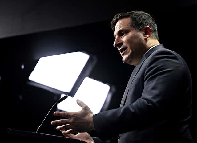 A man in profile with lights behind him gestures while he speaks.