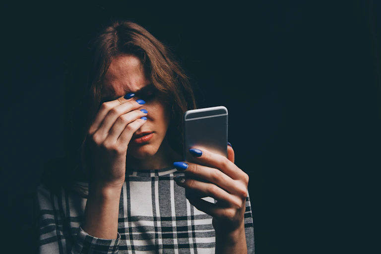 A young woman holds her mobile phone while anxiously resting her fingers on the bridge of her nose