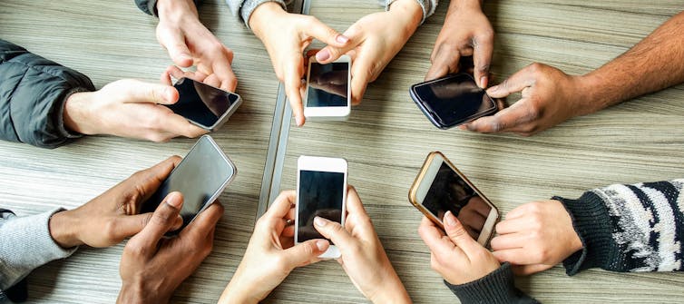 Overhead view of the hands of six people holding smartphones, seated around a table