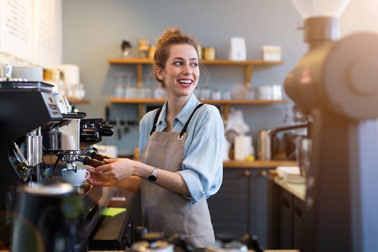 A young woman smiles wearing an apron as she uses a coffee machine to make a drink for a customer.