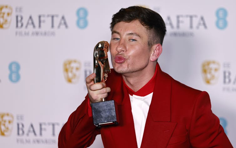 Barry Keoghan kisses his BAFTA award, which is shaped like a face. He wears a red suit.