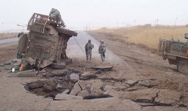 A vehicle on its side with two soldiers looking up at it.