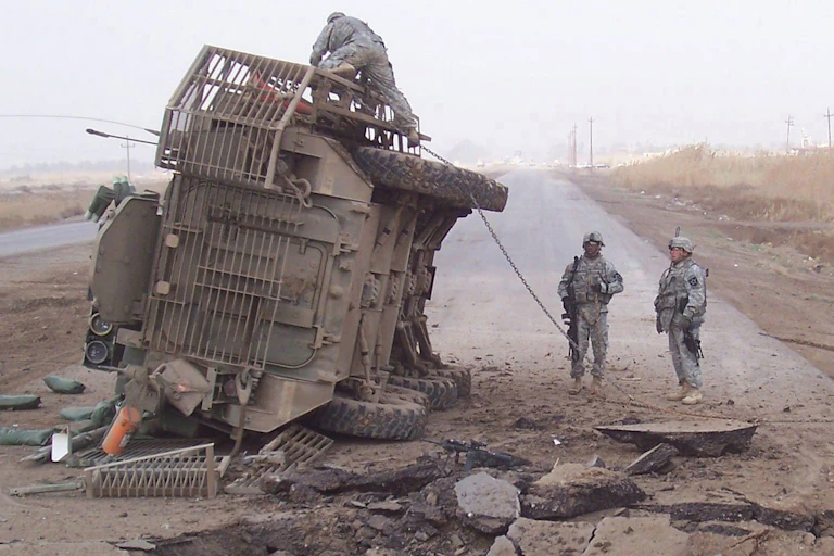 A vehicle on its side with two soldiers looking up at it.