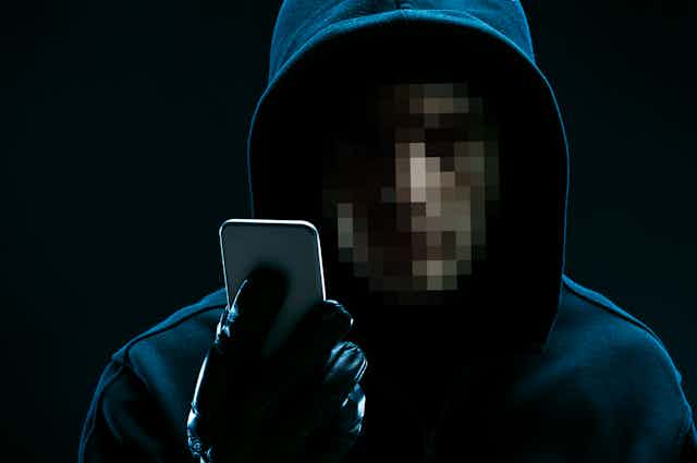 A hooded figure with a pixilated face holds a smart phone
