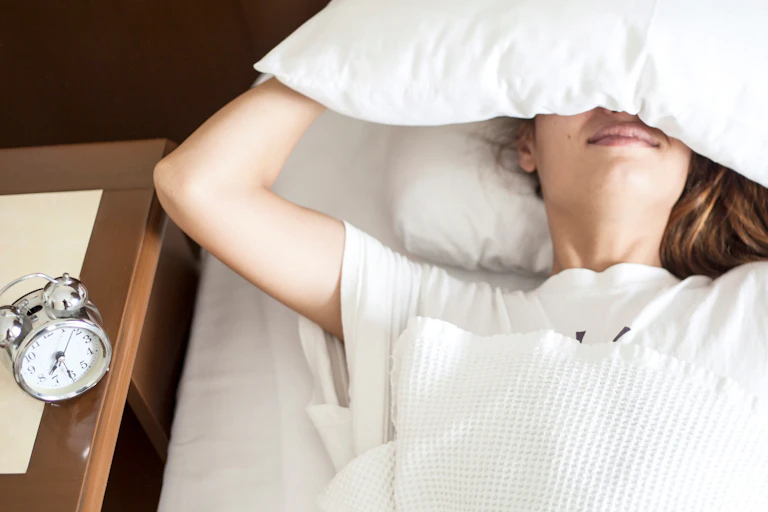 A woman lies in bed in a sunny room with a pillow over her eyes, an analog alarm clock on the bedside table reads 7:30