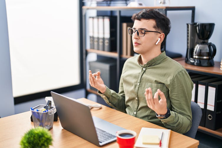 A young office worker with short hair and glasses wearing a green button up shirt sits at their desk with their eyes closed and airpods in, meditating with their hands in a meditation position, forefingers touching thumbs