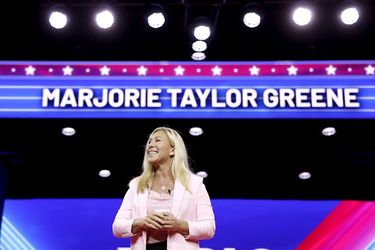 A blond woman in a pink jacket stands in front of many lights and a marquee that says 'Marjorie Taylor Greene'