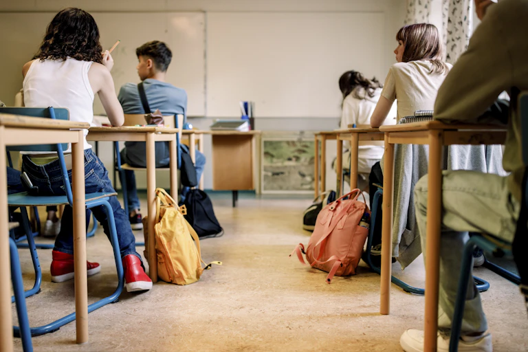 Students sit in desks and look to the left. The view is from behind.
