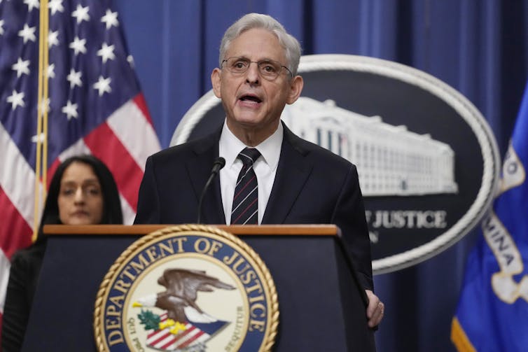 An older white man in glasses speaks from behind a podium with the U.S. Department of Justice logo on it