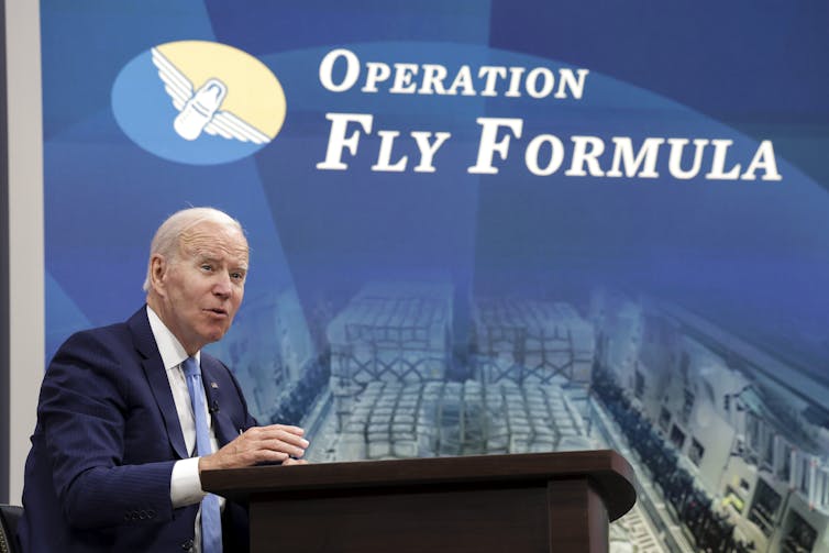 A formally dressed man with gray hair seated in front of a screen that says 'Operation Fly Formula'