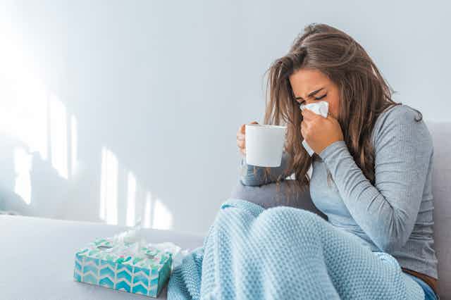 Young woman sitting on couch covered in a blanket, blowing her nose into a tissue and holding a cup of warm beverage