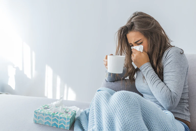 Young woman sitting on couch covered in a blanket, blowing her nose into a tissue and holding a cup of warm beverage