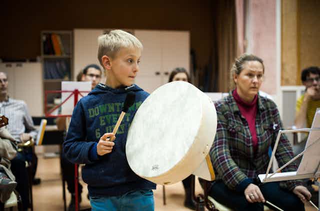 Un niño toca el tambor junto a otros adultos frente a atriles con partituras.