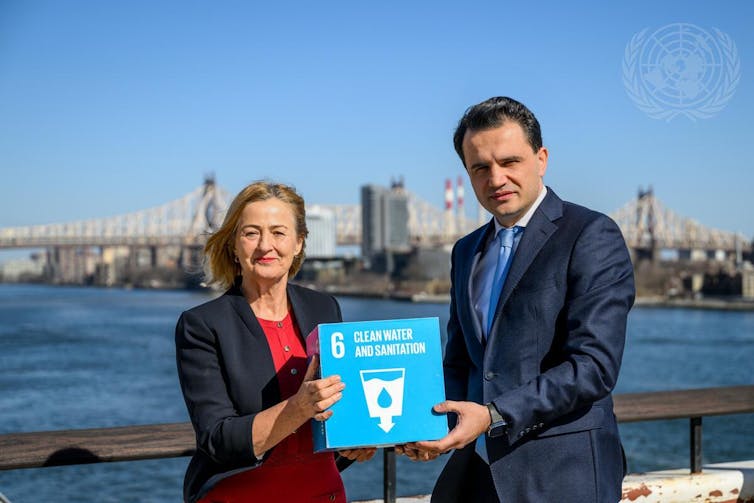 Yoka Brandt y Jonibek Ismoil Hikmat sostienen un cartel del ODS 6 con el río Este y la ciudad de Nueva York de fondo.