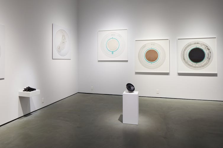 Seen on the wall, photographic images of circles, and a platform with a mound of lump-like black matter, and on the floor a black disc is seen sitting on a pedestal.