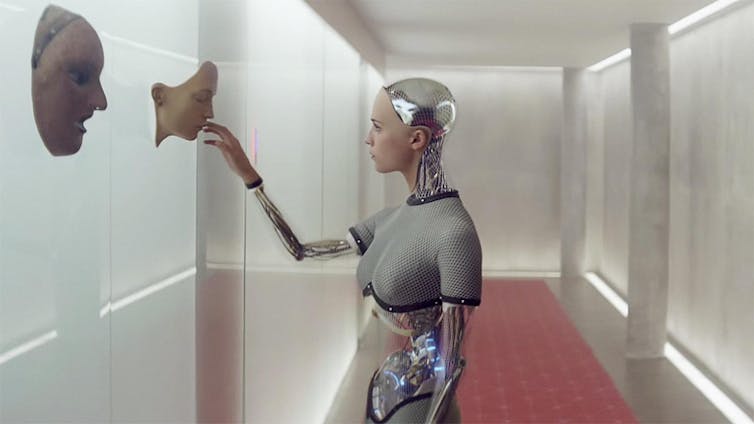 Una robot con apariencia femenina acaricia unas máscaras humanas en un pasillo blanco.