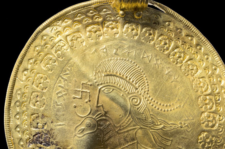 A gold circular pendant showing the head of a man and a horse, surrounded by runes.