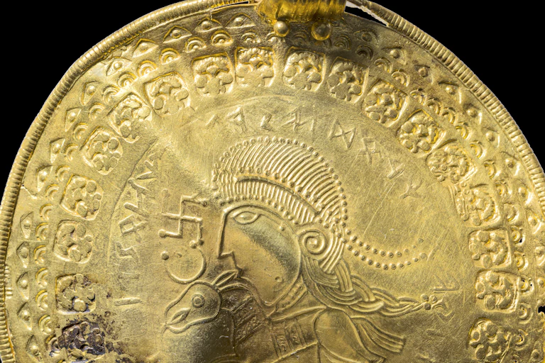 A gold circular pendant showing the head of a man and a horse, surrounded by runes.