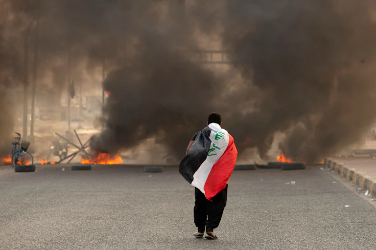A man is seen walking with a black, red and white flag draped over his back as he walks toward a billow of smoke.