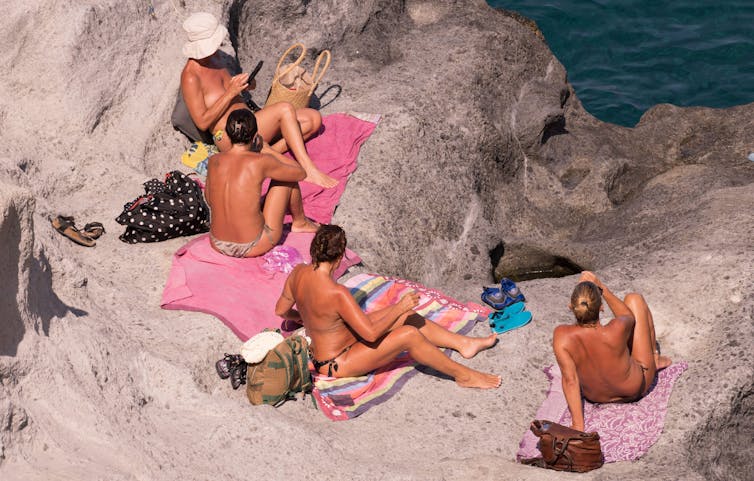 Women sit on rocks by the sea, sunbathing without tops on.