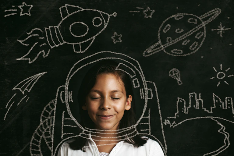 A young girl smiles against the backdrop of chalk drawings of a rocket, a plane, a planet and a parachute and a skyline.