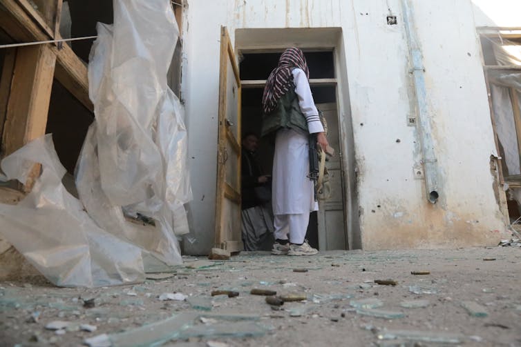Man wearing combat vest over traditional Afghan clothes and holding assault rifle looks through door of heavily damaged building