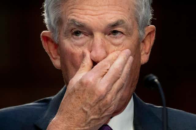 closeup of a white man with gray hair wearing a suit holding his right hand over his mouth