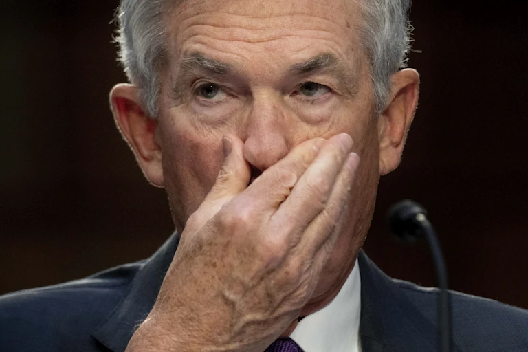 closeup of a white man with gray hair wearing a suit holding his right hand over his mouth