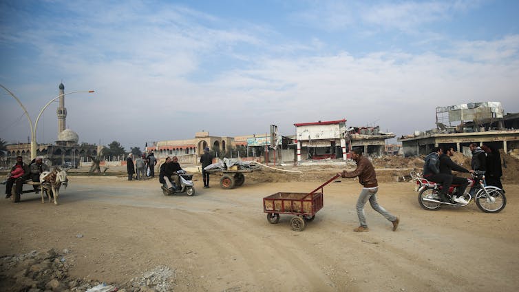 A man pushes a cart in a desolate looking area with sandy, dirt ground and blue skies.