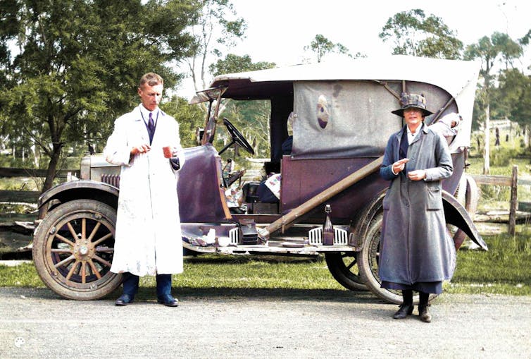 Father and daughter stand in front of a car in 1922. He is drinking from a cup. A telescope is strapped to the side of the car.