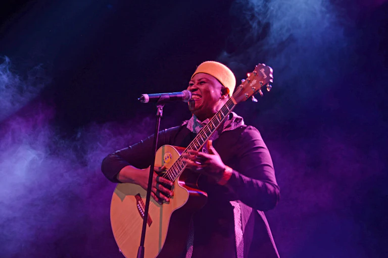 A woman smiles at a microphone, holding a guitar on a smoke-filled stage in a black dress and orange hat.