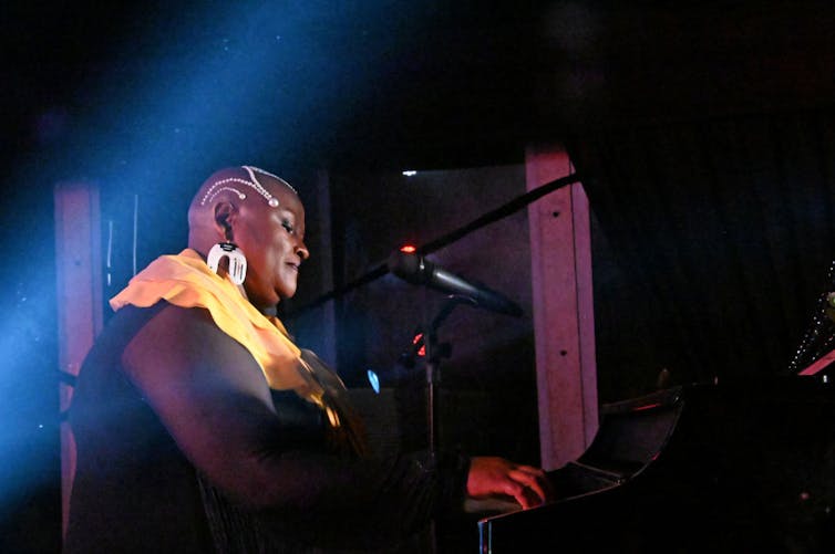 A woman on a dark stage plays the piano. She has gold decoration on her face and a bald head, looking serene.