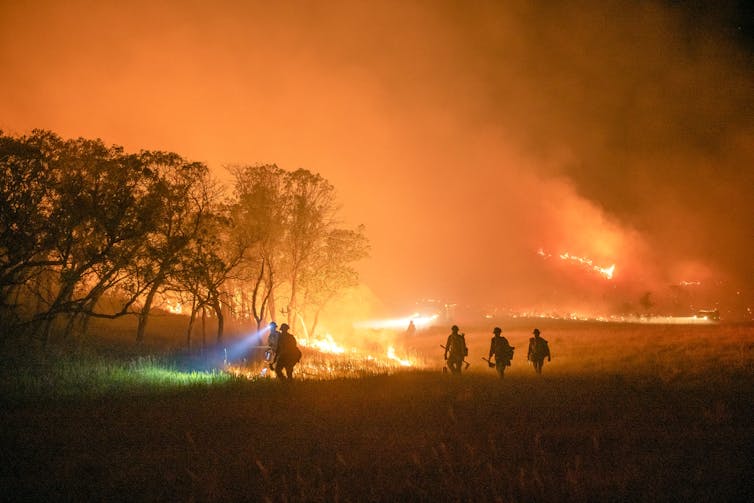 Silhouettes of five firefighters, one with a headlamp lighting the way, walking through a wooded field with fire burning in the background.