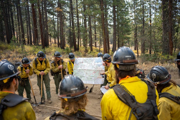 A dozen firefighters, some leaning on their Pulaski tools, look at a map of the fire. They're standing in a wooded area with tall pines behind them.