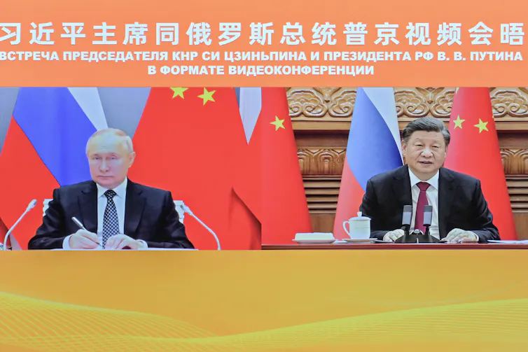 Two men sit in front of a row of mostly red flags and appear on a screen.