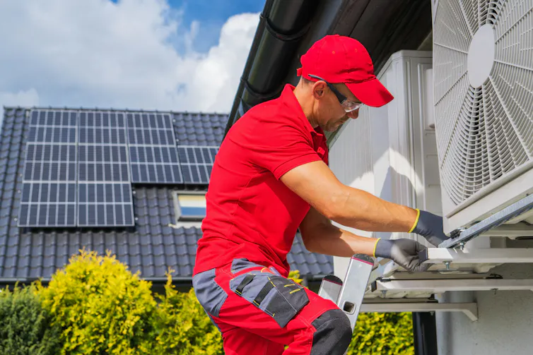 Worker performing heat pump maintenance, rooftop solar panels in the backround.