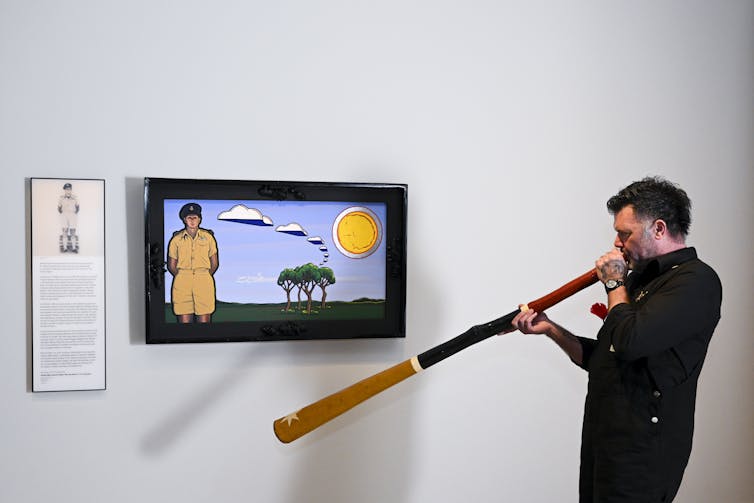 A man stands in front of a painting playing a yidaki.