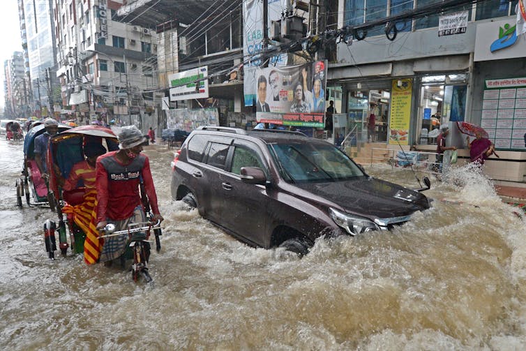 A pedicab driver looks over at an SUV making waves as both drive through knee-high water.