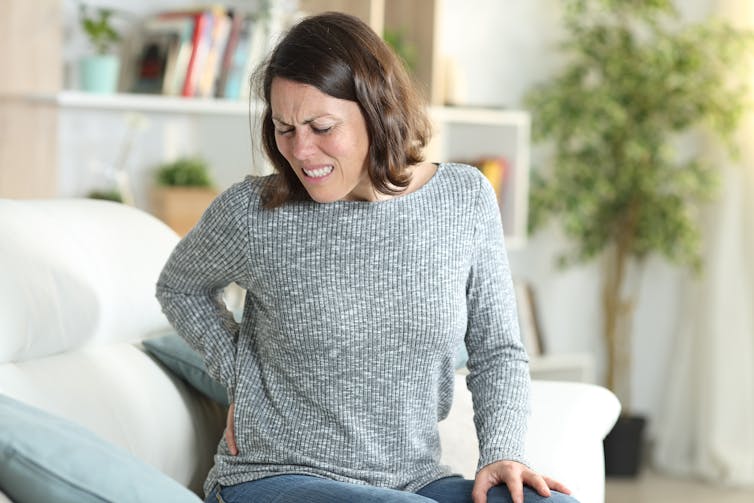 Woman sits on couch and grimaces with pain, rubs back